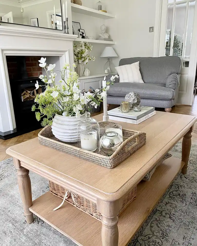 Light wood coffee table with woven tray, white flowers, glass candle holders, books, and wicker basket