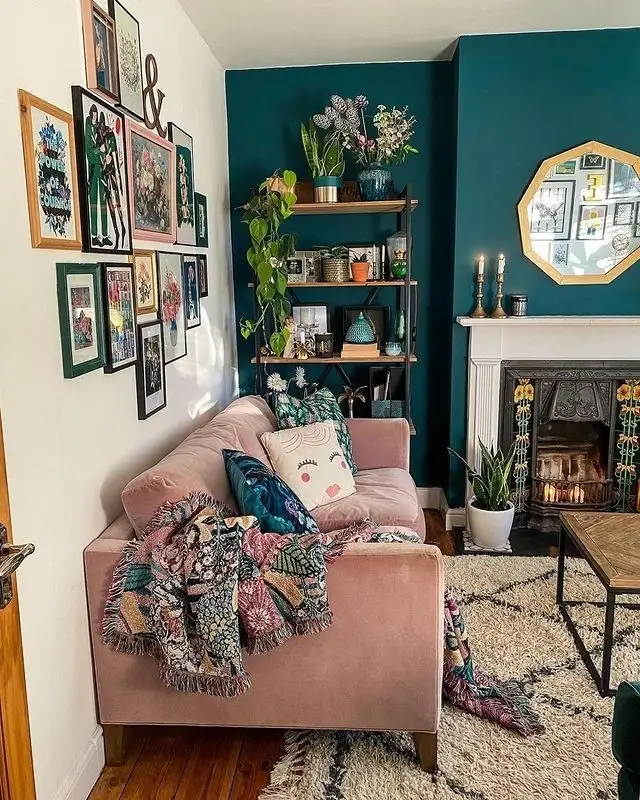 Living room with a deep teal fireplace wall, blush sofa, white fireplace surround, shelves, plants, and gallery art.