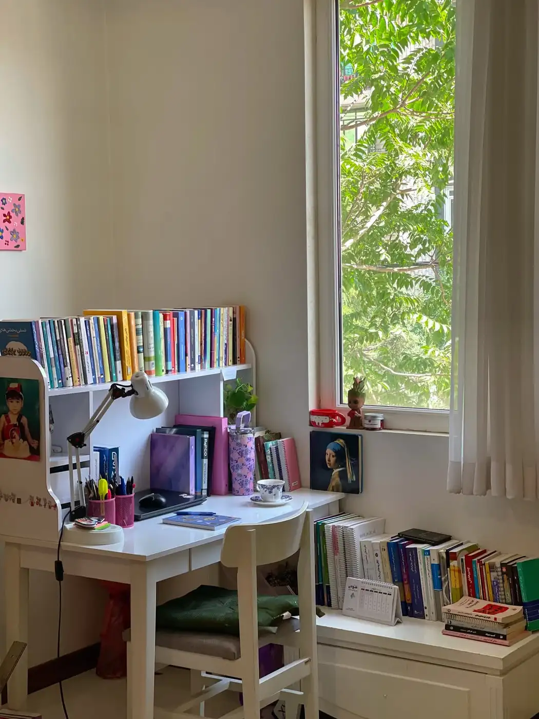 Small homework desk beside a window with laptop, books, lamp, pens, and low book storage