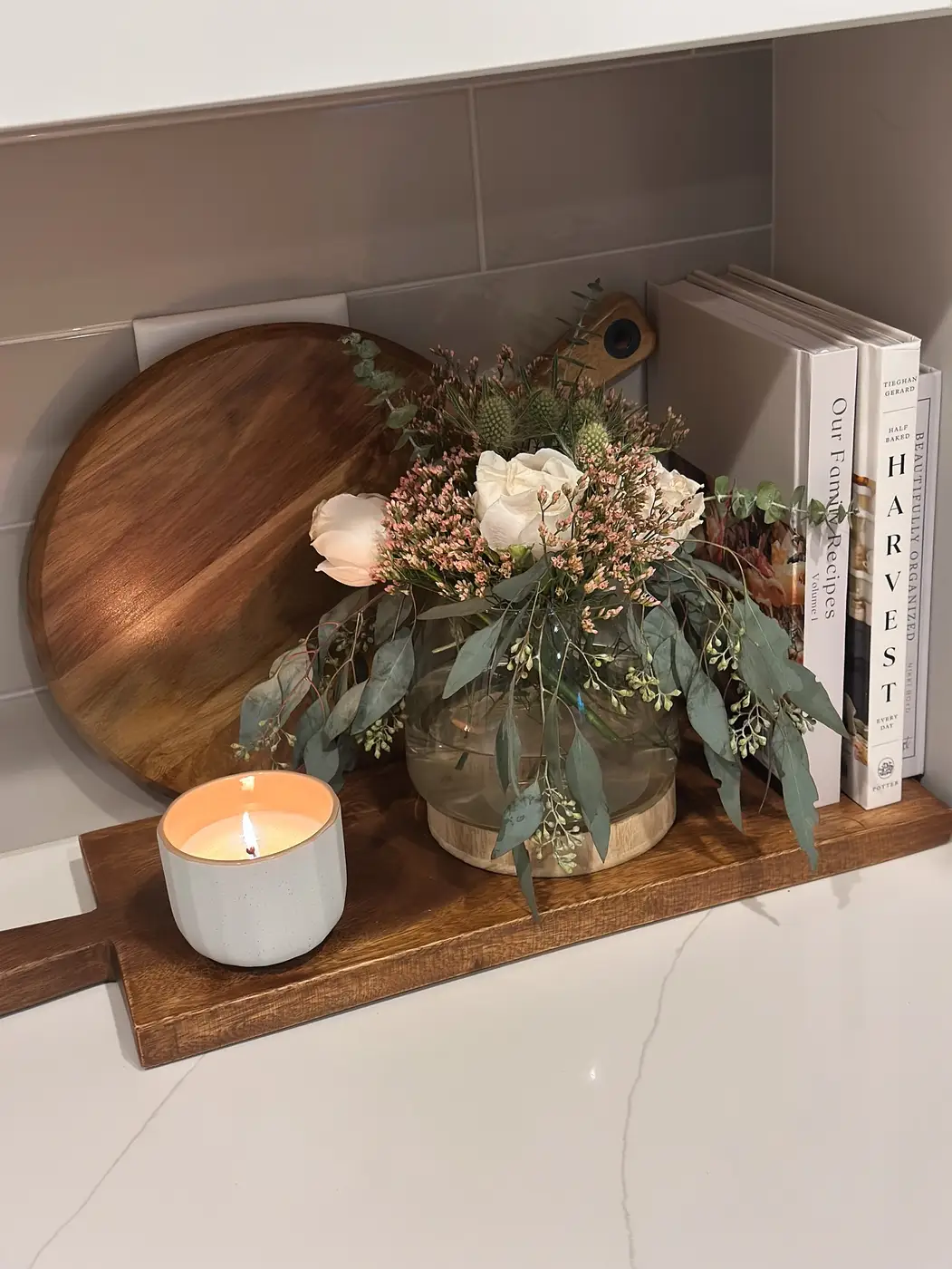 White kitchen counter with layered wooden boards, floral arrangement, cookbooks and lit candle
