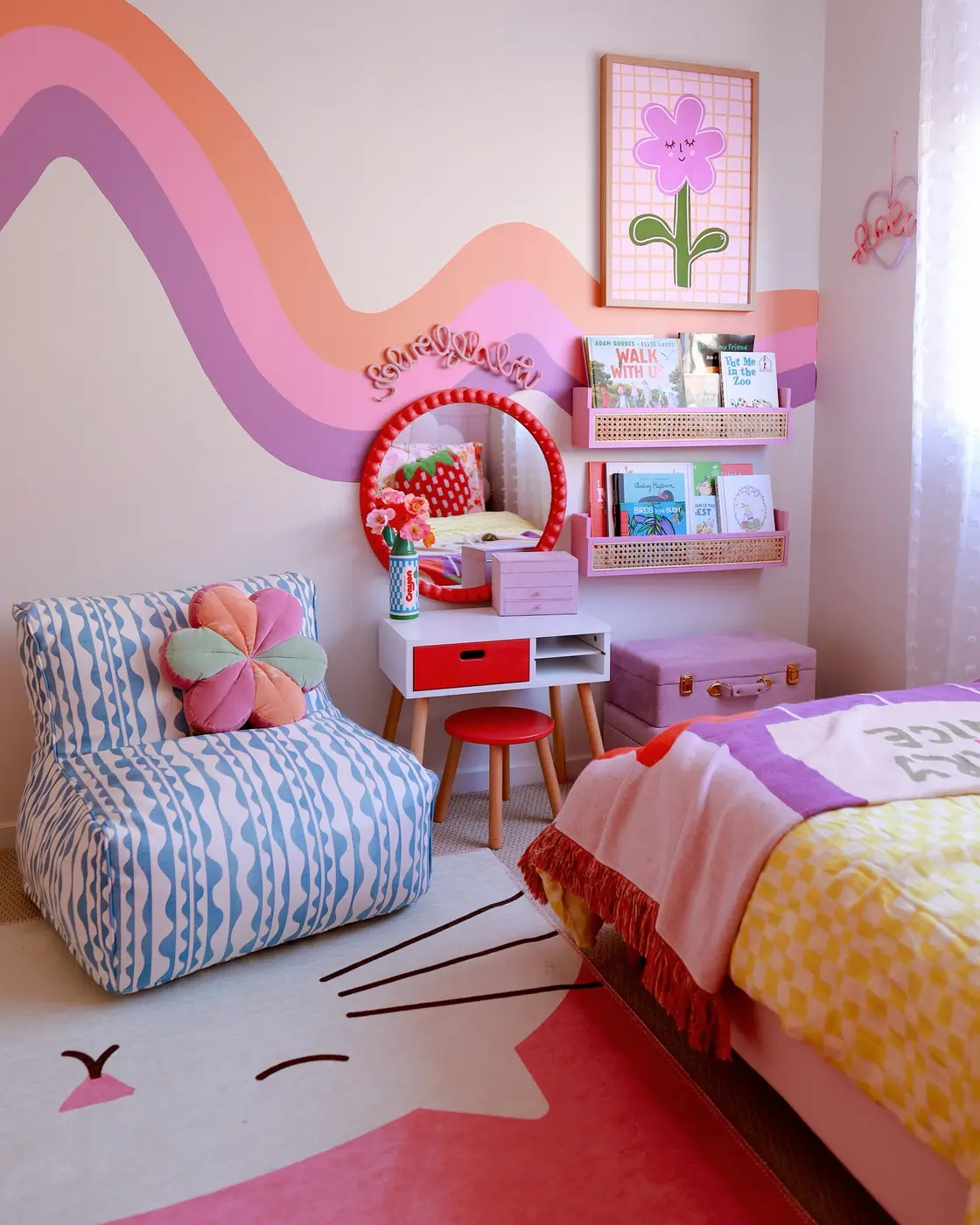 Colorful girls bedroom with large painted rainbow wall, bookshelves, mirror, patterned bedding, and flower cushion