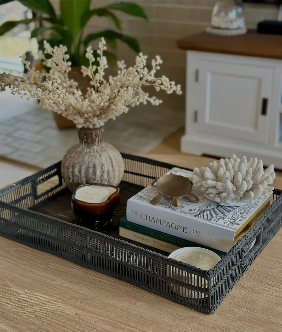 Dark woven coffee table tray with white coral decor, stacked books, weathered vase, dried stems, and amber candle