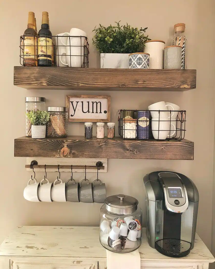 Rustic farmhouse coffee bar with dark wood shelves, wire baskets, hanging mugs, syrup bottles, coffee machine, and pod jar
