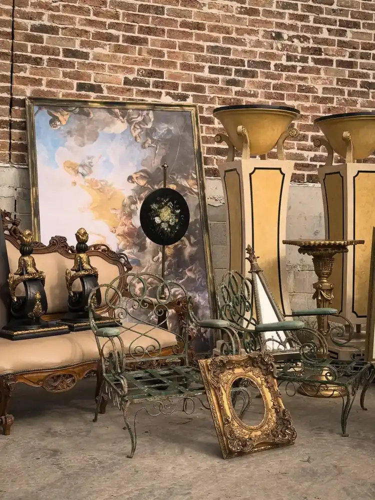 Vintage seating area with worn green metal chairs, gilded frame, carved furniture, and classical artwork