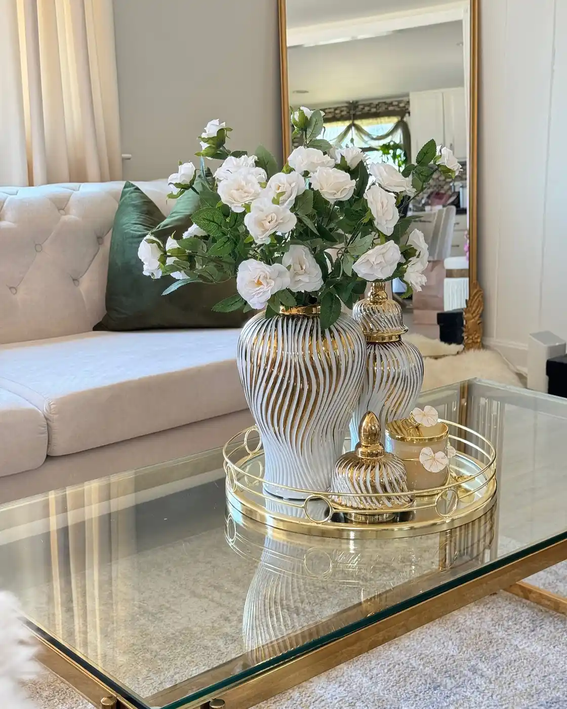 Glass coffee table decorated with gold tray, striped gold vase, white roses, candle, and lidded jar