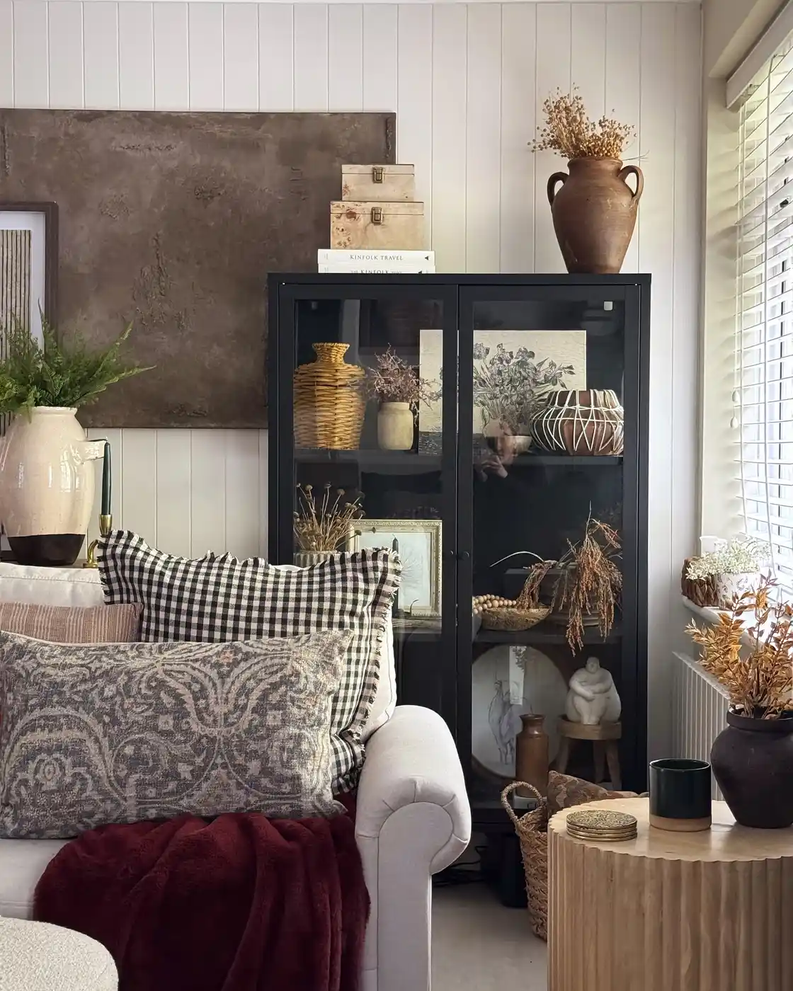 Tall black glass display cabinet filled with baskets, pottery, dried stems, framed art, and neutral decor