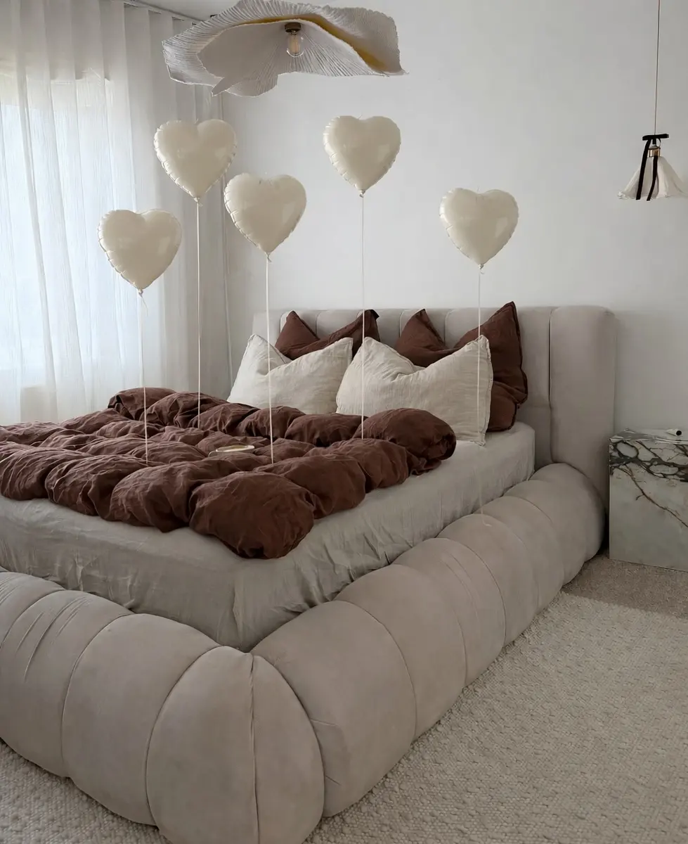 Cream upholstered bedroom with deep brown bedding, white pillows, heart-shaped balloons, and sculptural ceiling light.