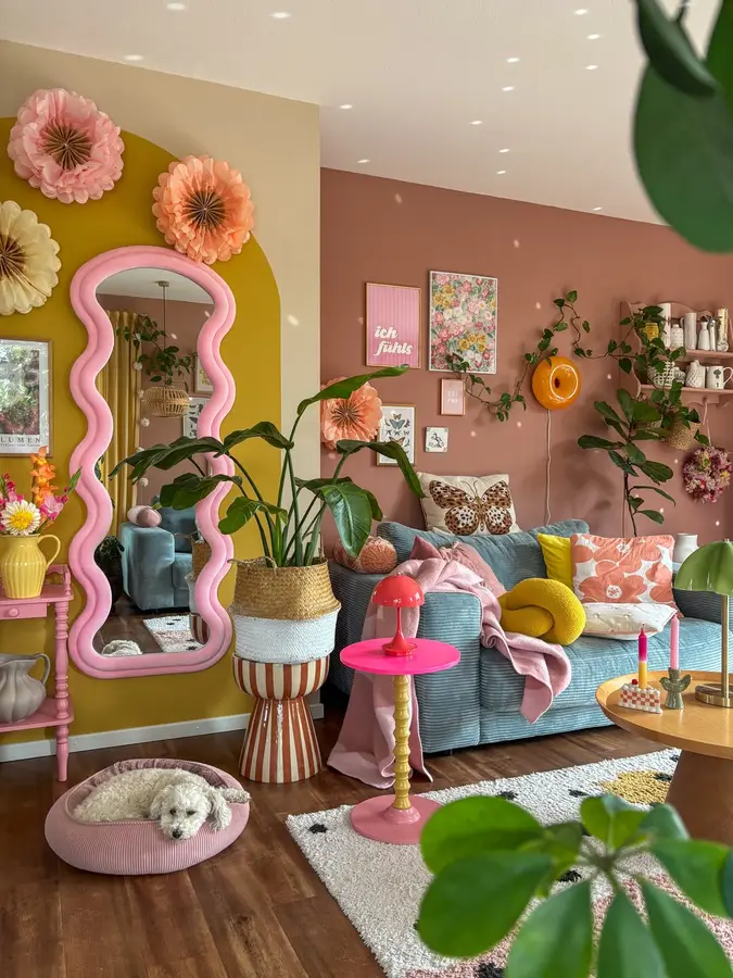 Colorful living room with oversized wavy pink mirror, mustard wall, pink side table, blue seating, and green plants