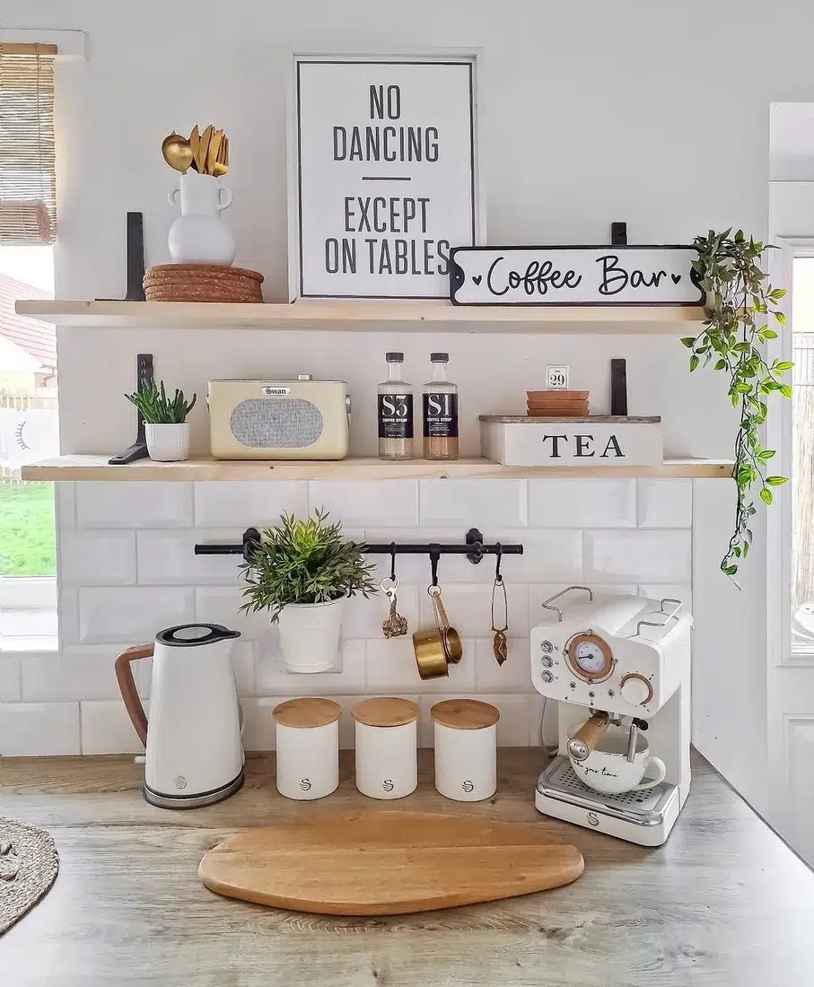 Modern farmhouse coffee bar with white tile, pale wood shelves, cream appliances, black rail, canisters, and simple neutral decor