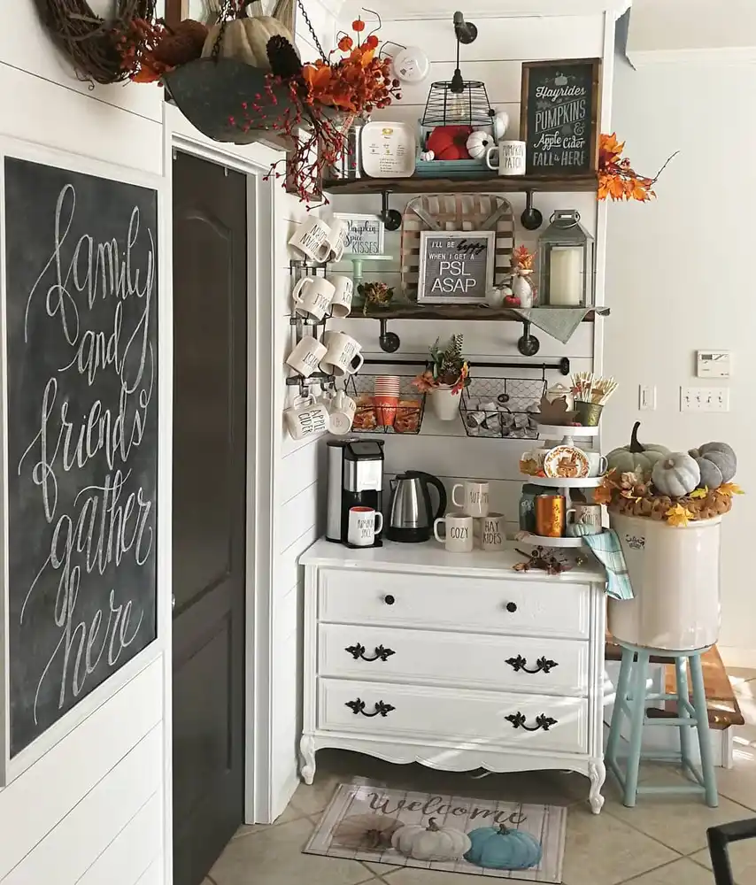 Fall coffee station on a white cabinet with wall shelves, hanging mugs, wire baskets, pumpkins, and autumn signs