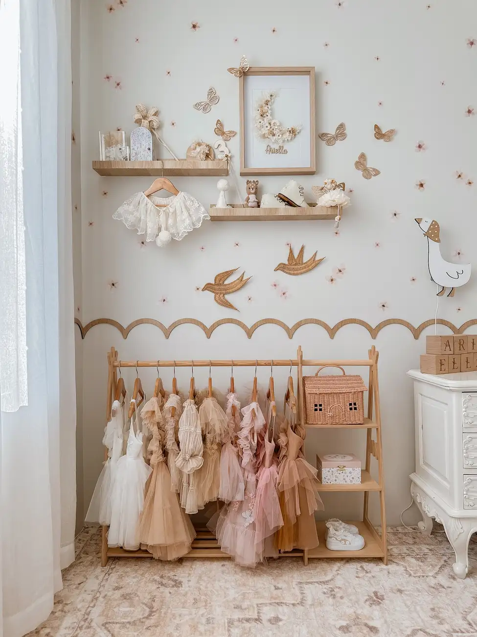 Girls dress-up corner with wooden clothing rail, blush dresses, small shelves, baskets, and butterfly wall decor