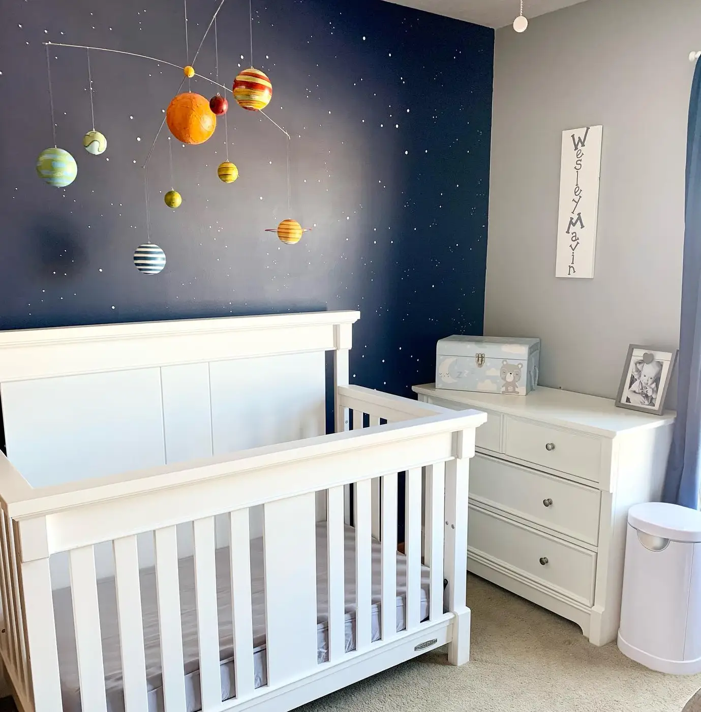 Space nursery with hanging planet mobile, dark blue wall, white crib, and white storage furniture