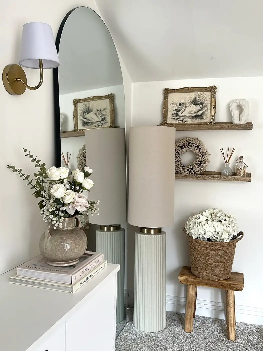 Small sloped-ceiling corner with tall arched mirror, floor lamp, floating shelves, flowers, and rustic stool