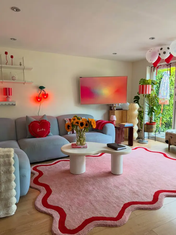 Playful living room with pale blue sofa, pink rug with red wavy border, red heart cushion, striped lamp, and wall light