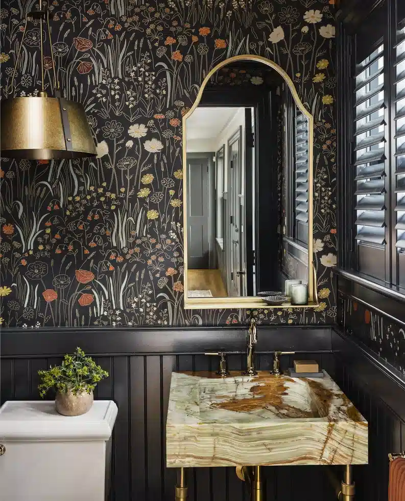 Moody bathroom with black floral wallpaper, black paneling, arched brass mirror, and green stone sink