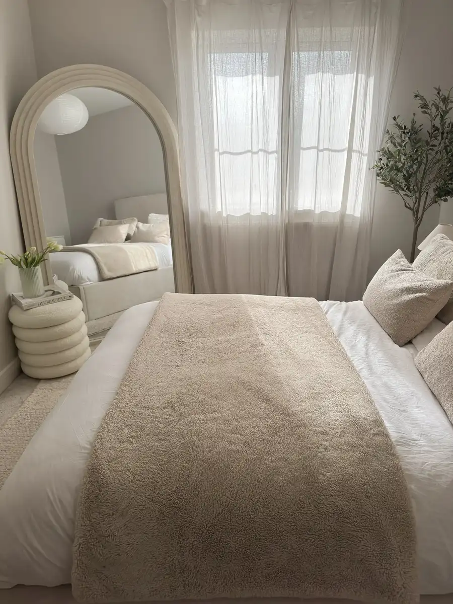 Light cream bedroom with large arched mirror, sheer curtains, textured bedding, rounded side table, and leafy indoor tree.
