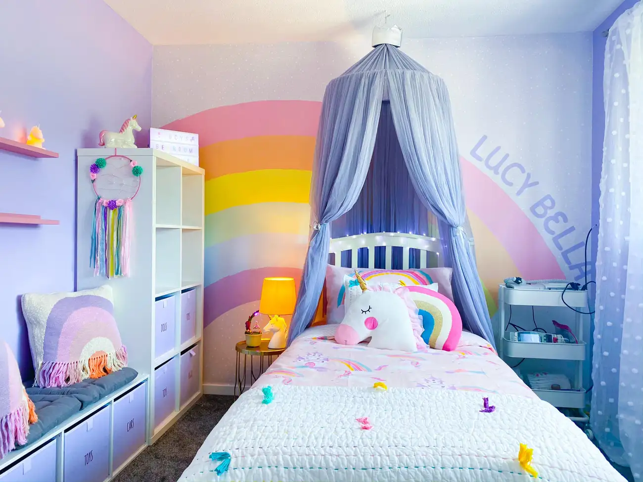 Lavender girls bedroom with painted rainbow wall, soft blue canopy, low toy storage, and colorful bedding