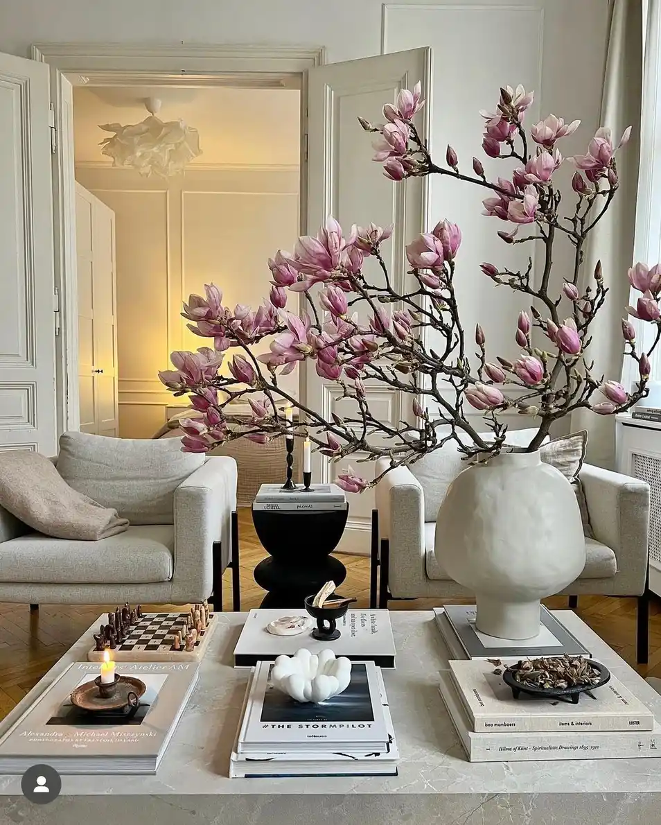 Large coffee table styled with oversized magnolia branches, stacked books, chess set, candle, and bowls