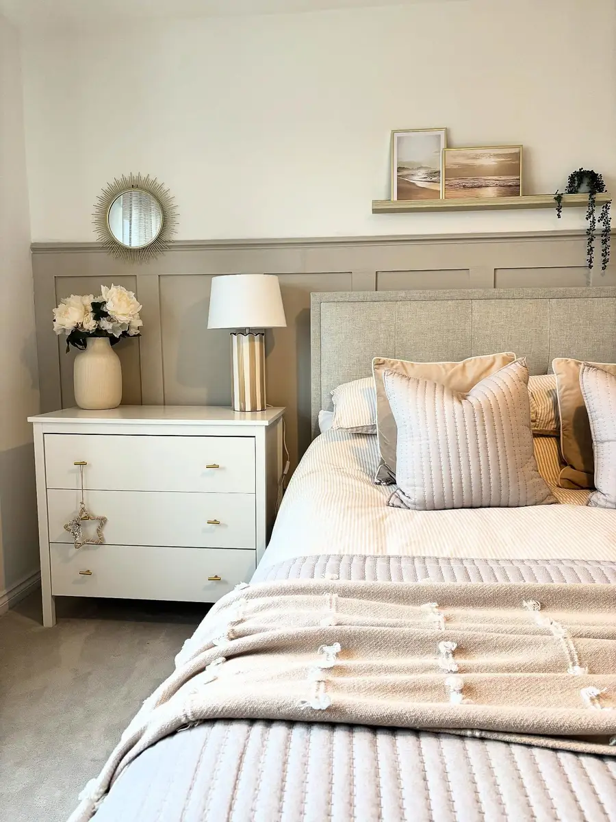 Beige bedroom with white dresser, paneled wall, upholstered headboard, layered neutral bedding, picture ledge, and sunburst mirror.