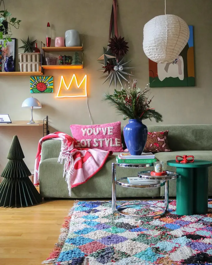 Colorful living room with olive sofa, pink throw, blue vase, bright side table, patterned rug, and neon wall light