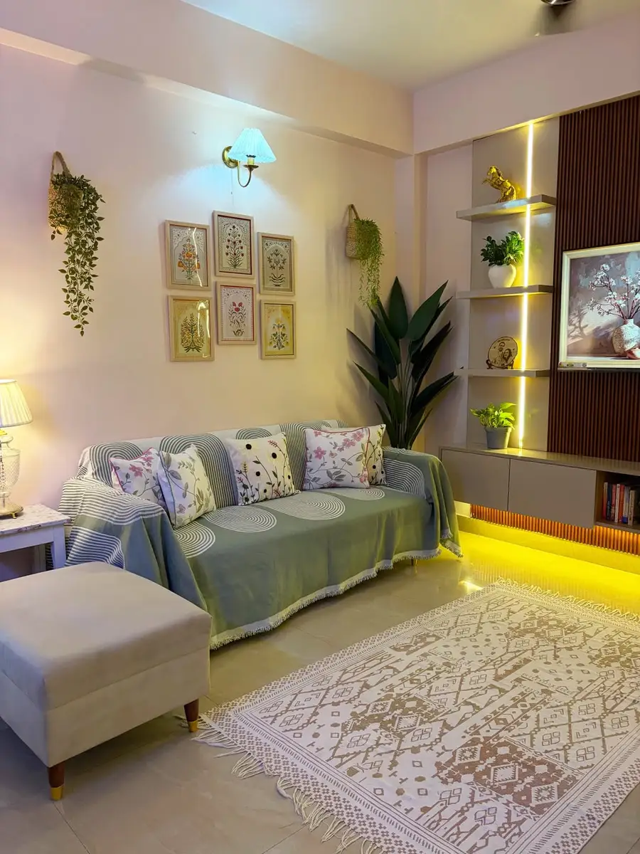 Small living room with warm-lit built-in shelves, indoor plants, botanical prints, and a sofa with floral cushions.