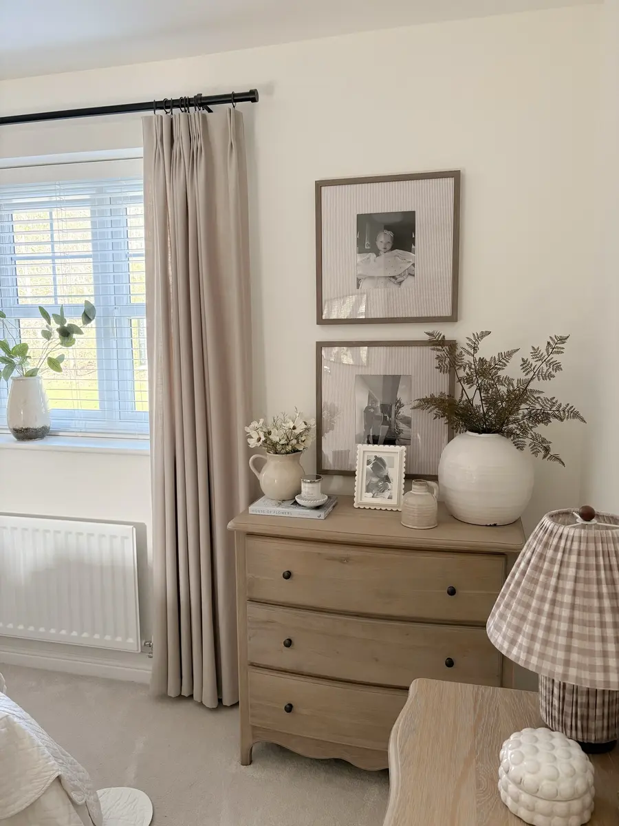Warm neutral dresser corner with pale wood drawers, framed photos, cream ceramics, taupe curtains, leafy stems, and gingham lamp.