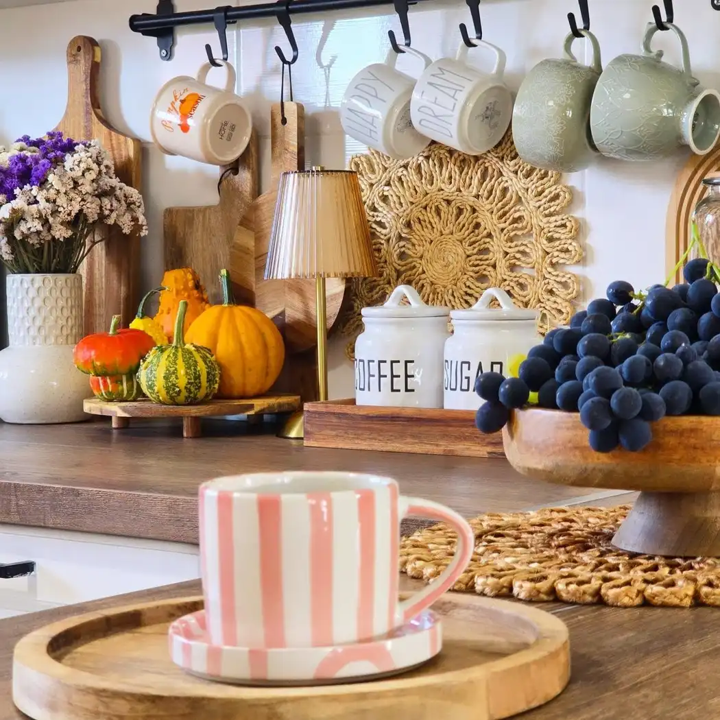 Cozy kitchen counter with hanging mugs, wooden cutting boards, pumpkins, lamp, canisters and bowl of grapes
