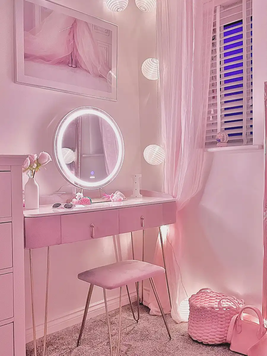 Small pink vanity with round lighted mirror, tucked stool, woven basket, and soft curtains