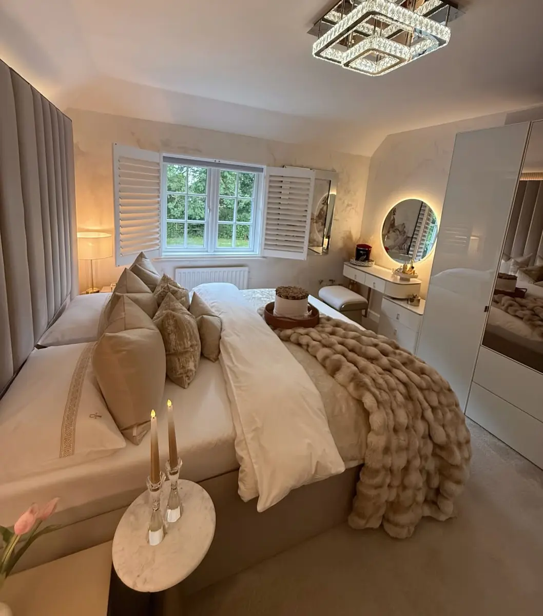 Luxurious taupe bedroom with layered pillows, plush throw, glossy wardrobes, round lighted mirror, shutters, and modern ceiling light.