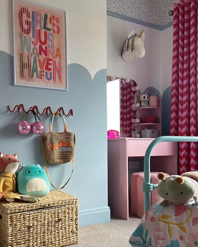 Colorful girls bedroom with blue walls, pink vanity, red curtains, wall hooks, woven trunk, and playful decor