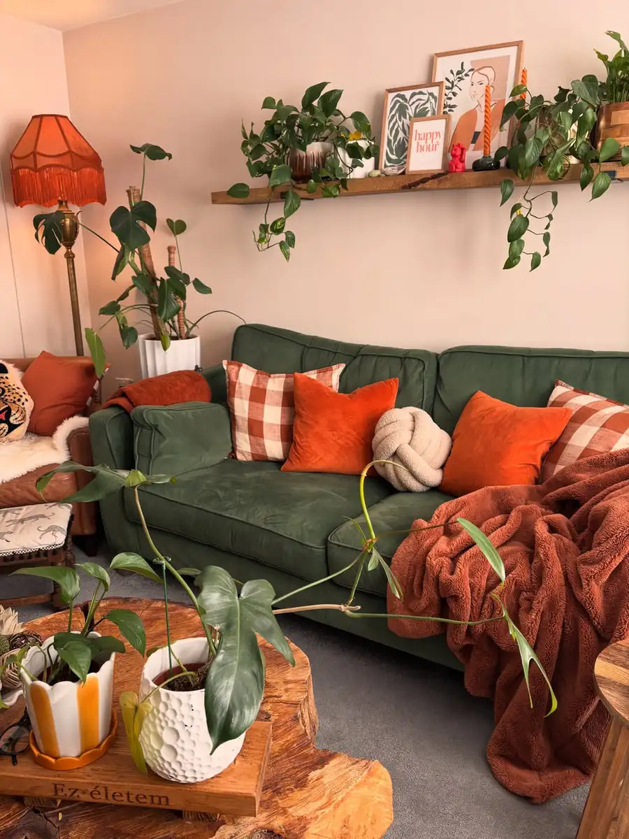 Cozy living room with a deep green sofa, burnt orange pillows and throw, wood coffee table, trailing plants, and warm lamp.