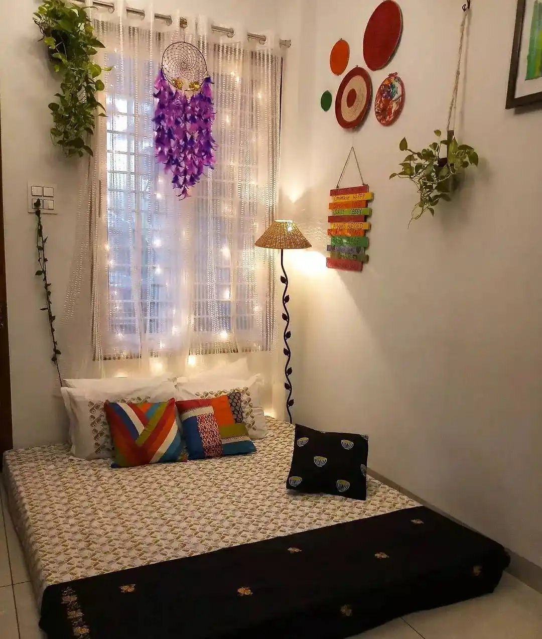 Low floor daybed in a living room corner with colorful cushions, hanging plants, fairy lights, and wall decor