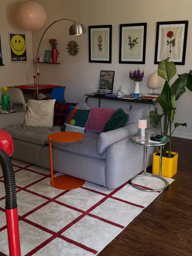 Neutral gray living room with orange side table, yellow planter, green cushions, colorful rug, plants, and bright accents