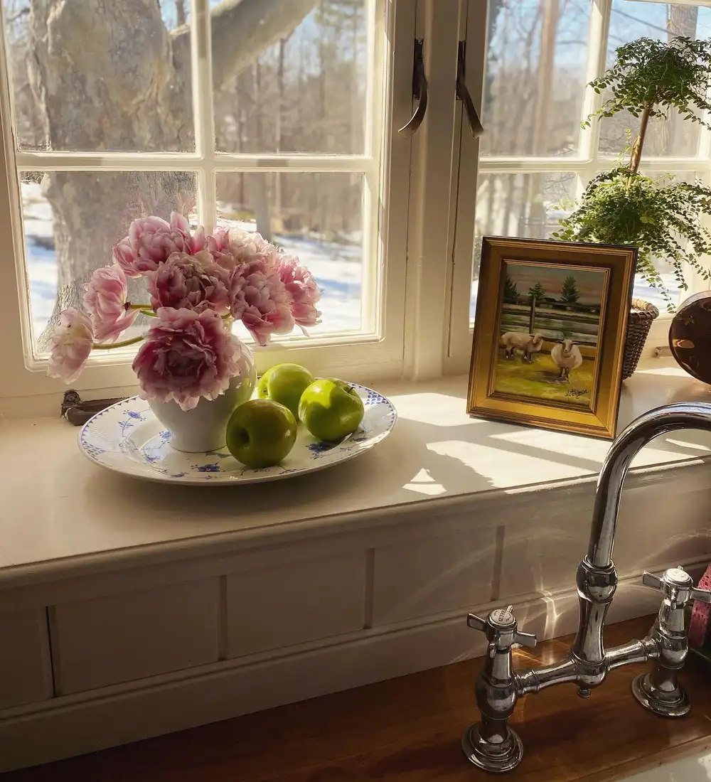 Kitchen window sill with pink flowers, green apples, patterned platter, and small framed picture