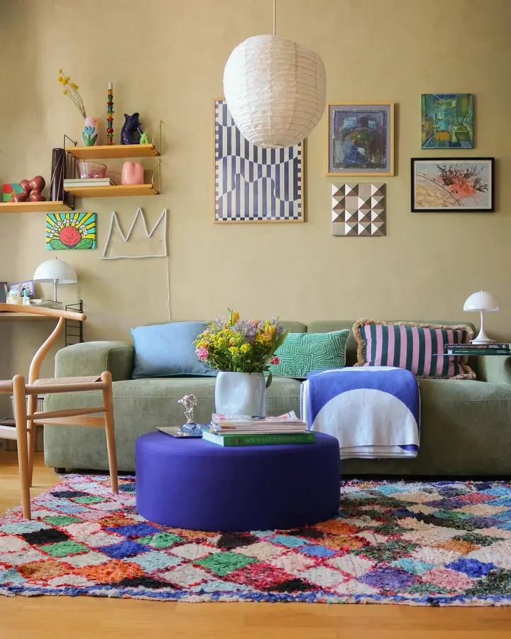Colorful living room with olive sofa, deep purple ottoman, patterned cushions, multicolor rug, pale blue accents, and plants