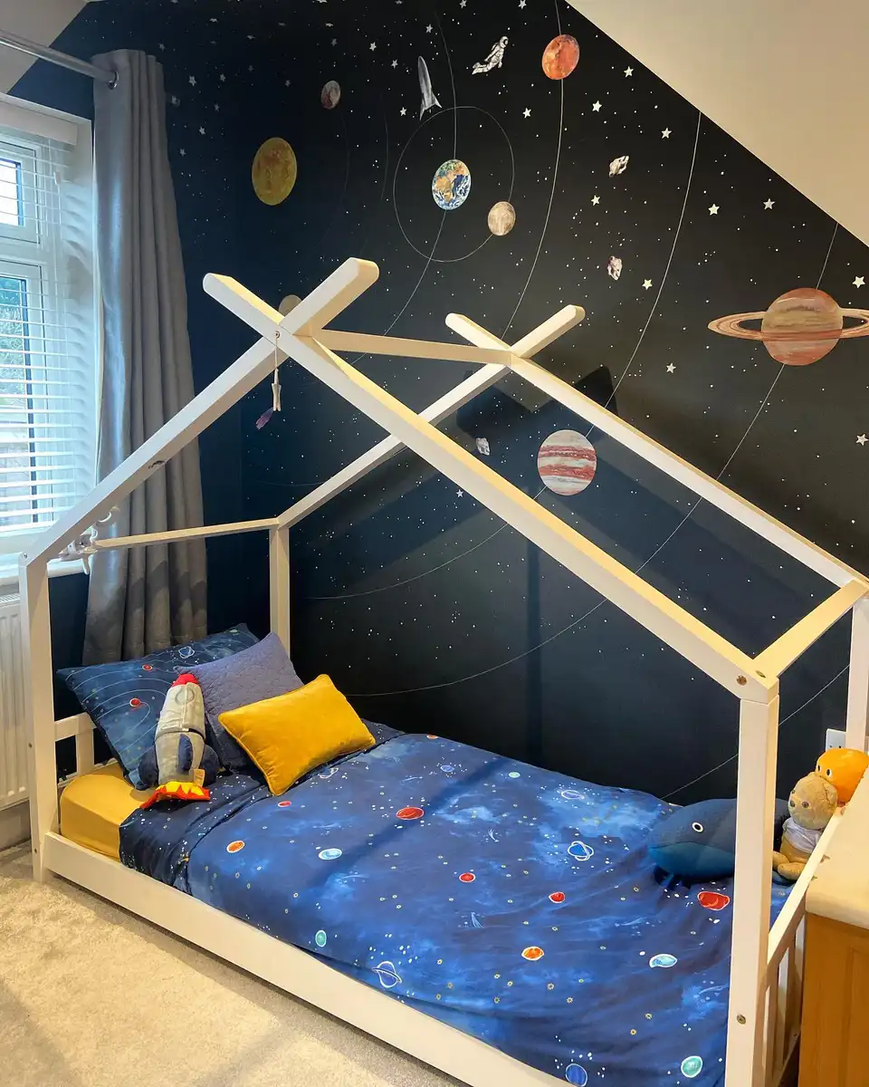 Space-themed bedroom with sloped dark wall, planets, astronauts, rocket graphics, white bed, and blue bedding