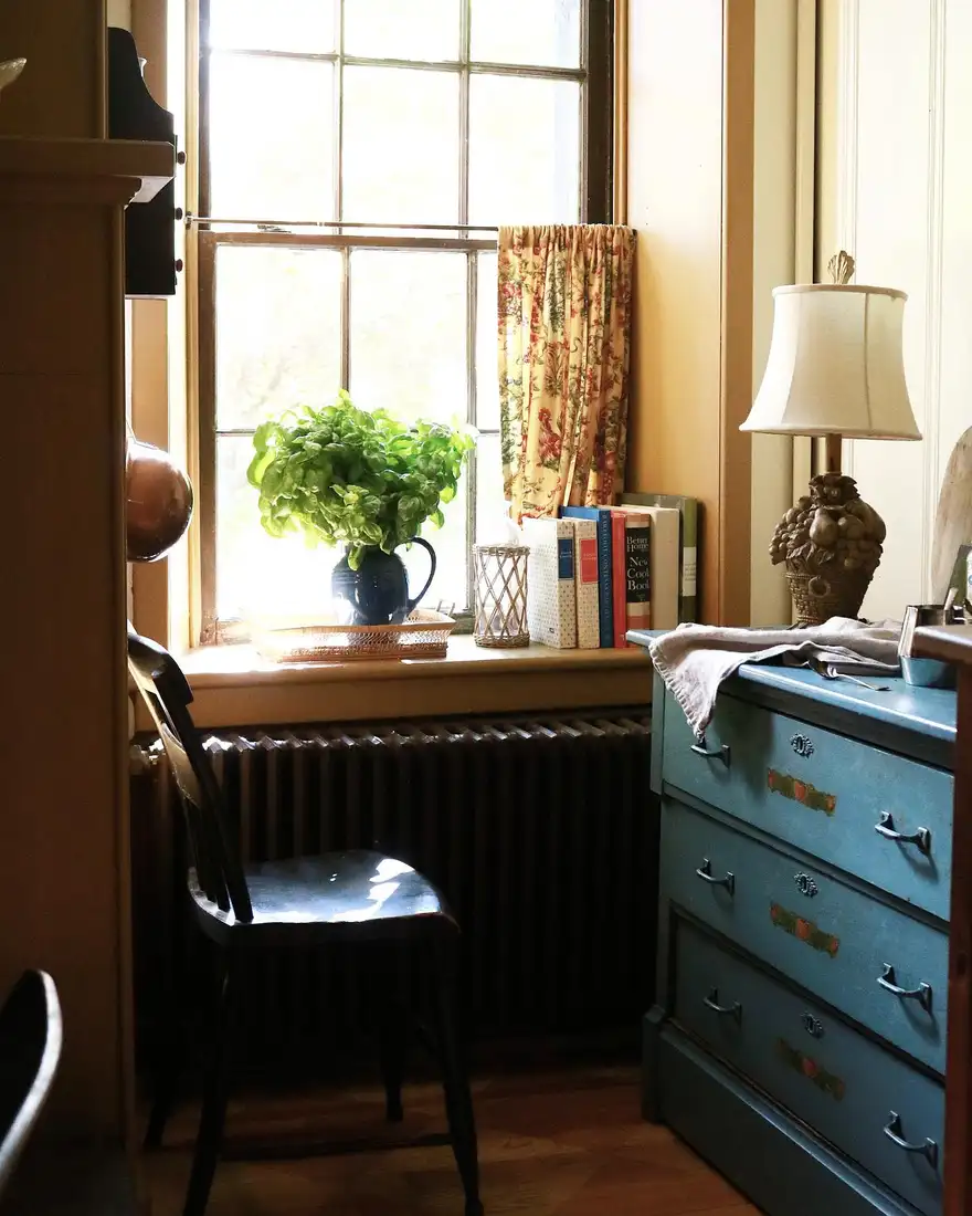 Deep kitchen window sill styled with cookbooks, leafy greenery in a dark pitcher, and candle holder
