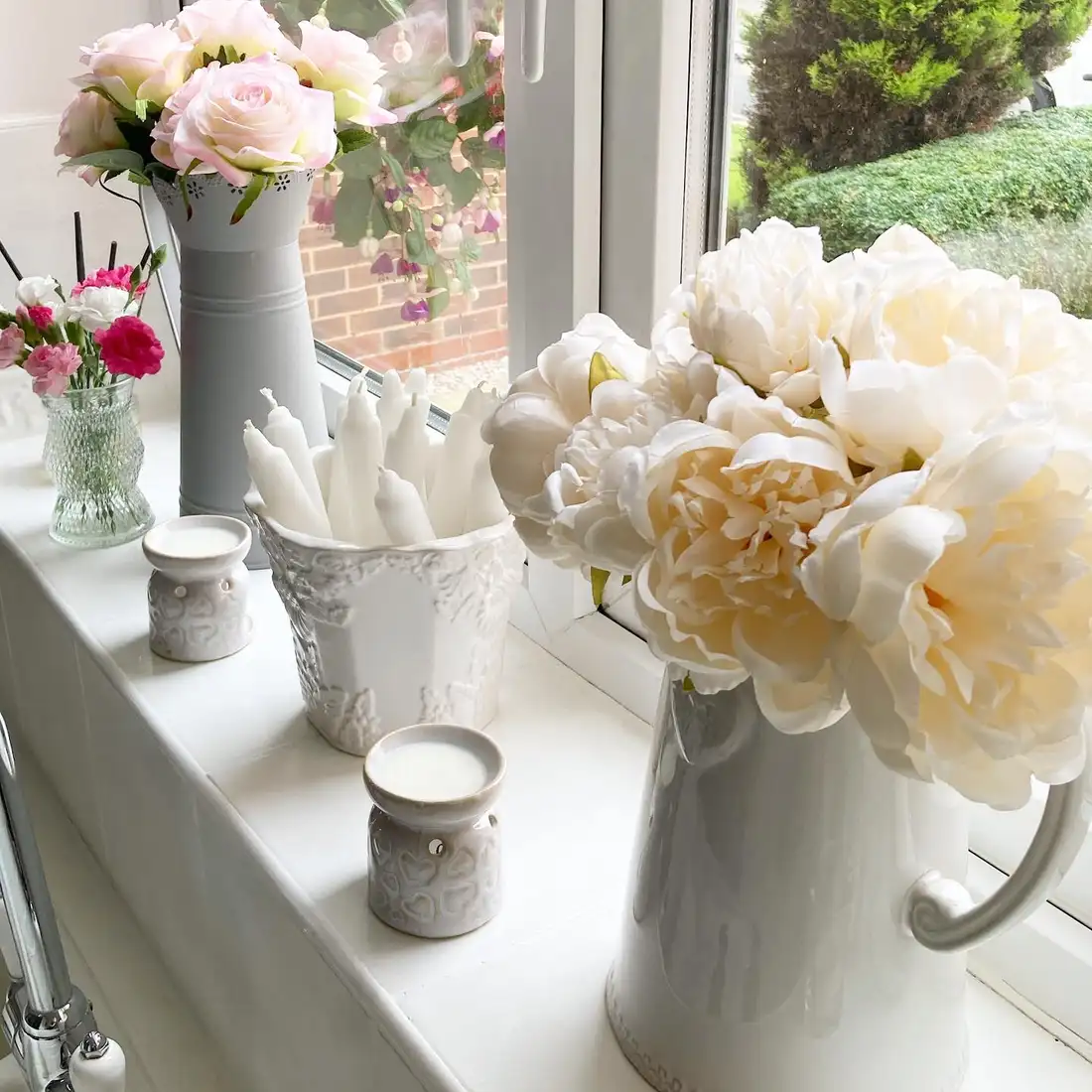 White kitchen window sill display with peonies, roses, white pitchers, candles, and ceramics