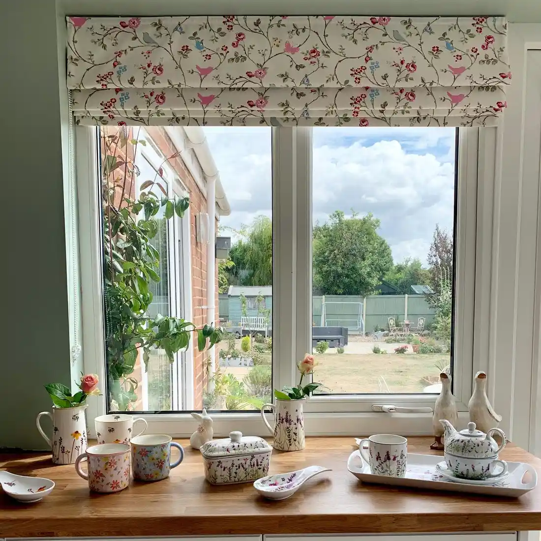 Wide kitchen window sill styled with floral mugs, teapot, pitchers, trays, and small flowers