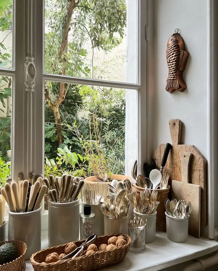 Kitchen window sill used for utensil storage with ceramic crocks, wooden boards, and small plant
