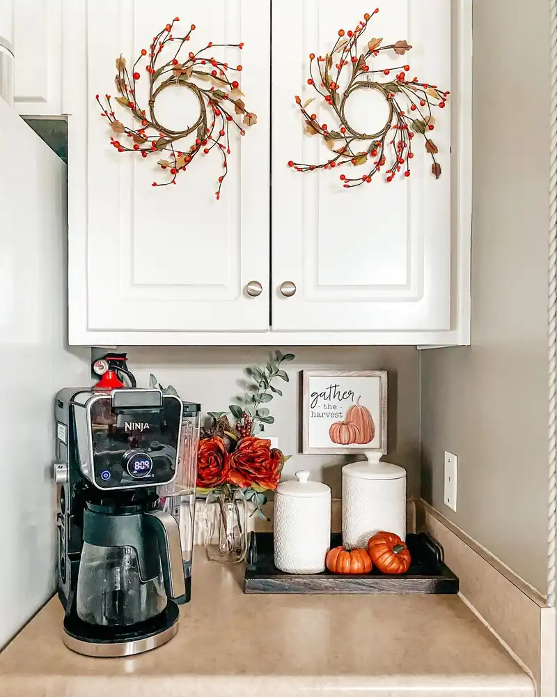 Fall coffee station beneath white cabinets with small wreaths, orange flowers, white canisters, and a pumpkin print