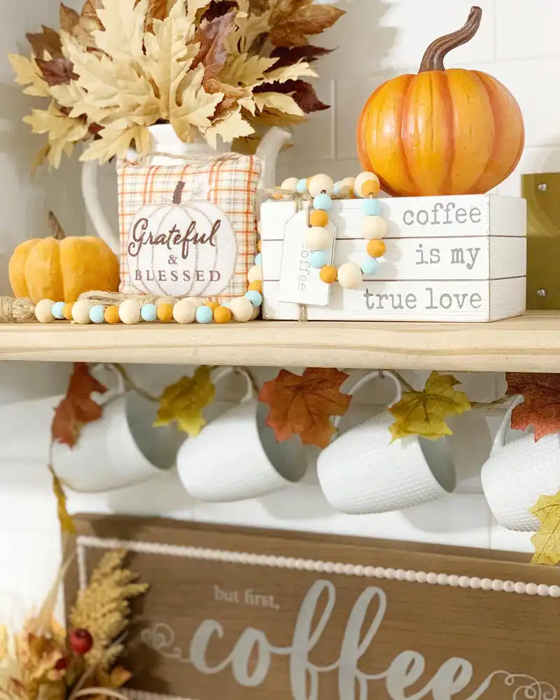 Neutral fall coffee station with white hanging mugs, orange leaf garland, pumpkins, dried stems, and wooden beads