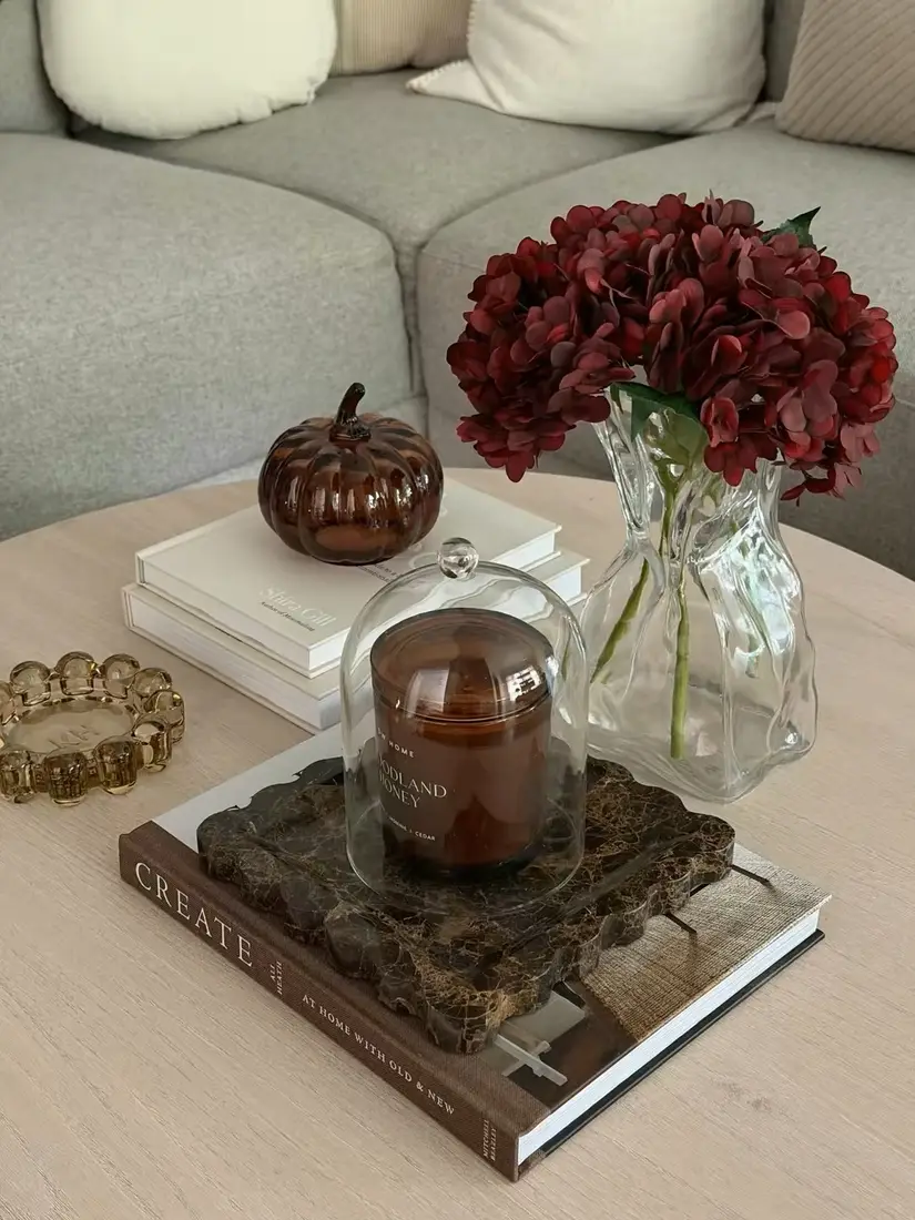 Coffee table decorated with burgundy flowers in a clear vase, amber candle, brown glass pumpkin, and stacked neutral books