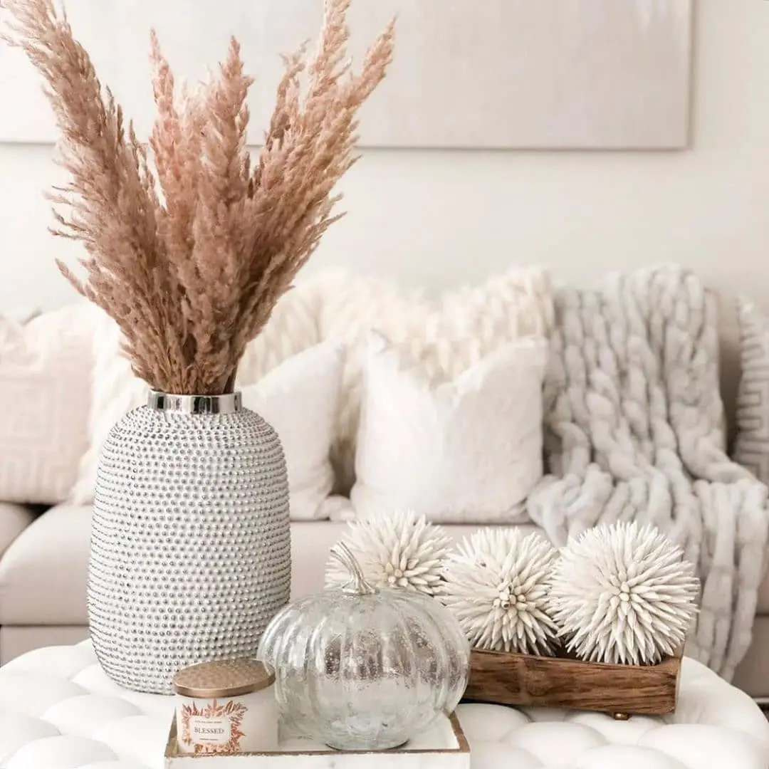Neutral fall coffee table decor with feathery dried stems, textured silver vase, pale glass pumpkin, candle, and wooden tray