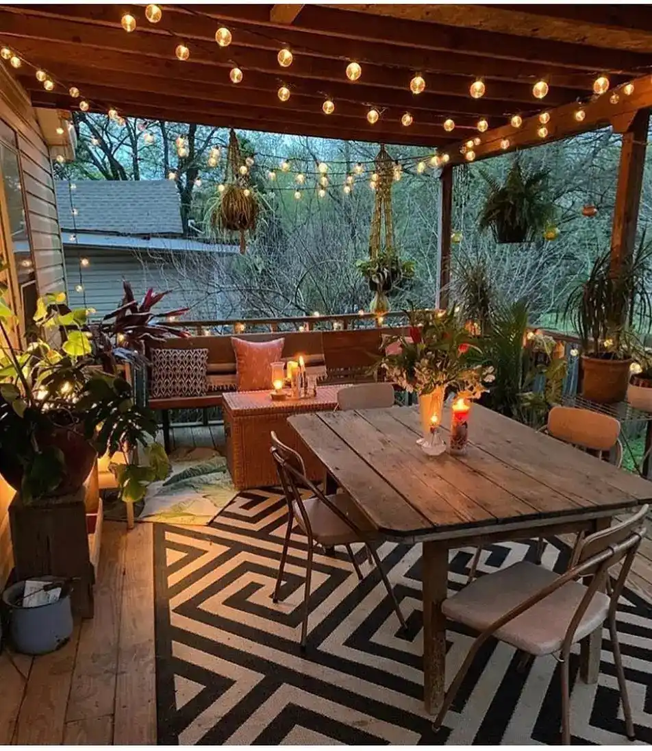Covered back porch with a long wood dining table, patterned rug, string lights, and potted plants.