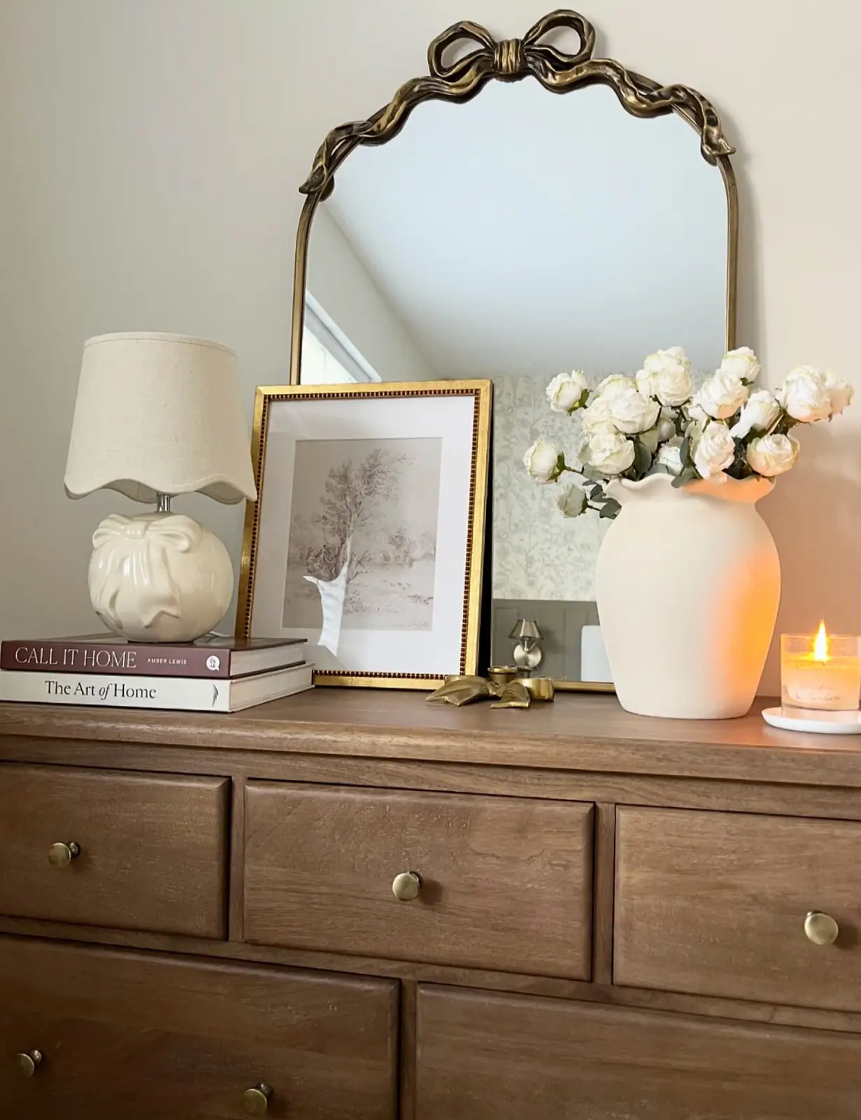 Wood dresser styled with ornate mirror, leaning framed art, table lamp, books, and white flowers