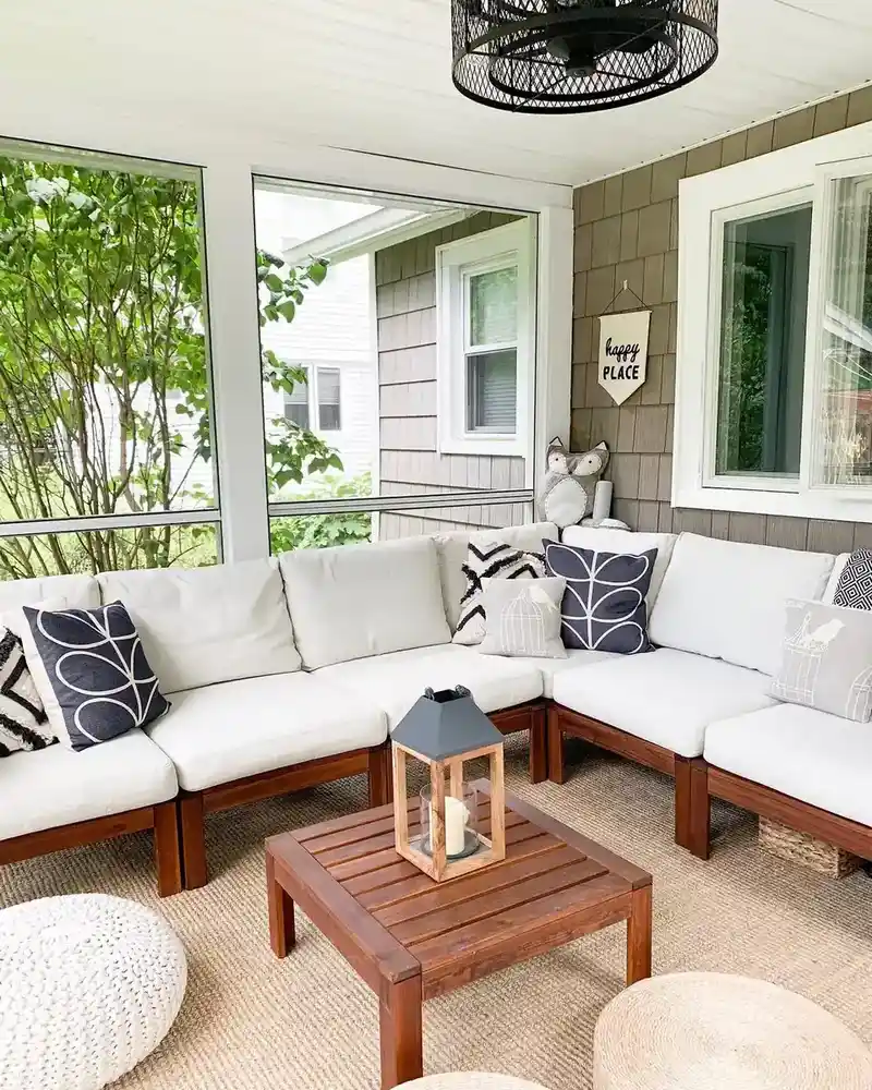 Bright covered porch with a pale sectional sofa, patterned pillows, wood coffee table, and large windows.
