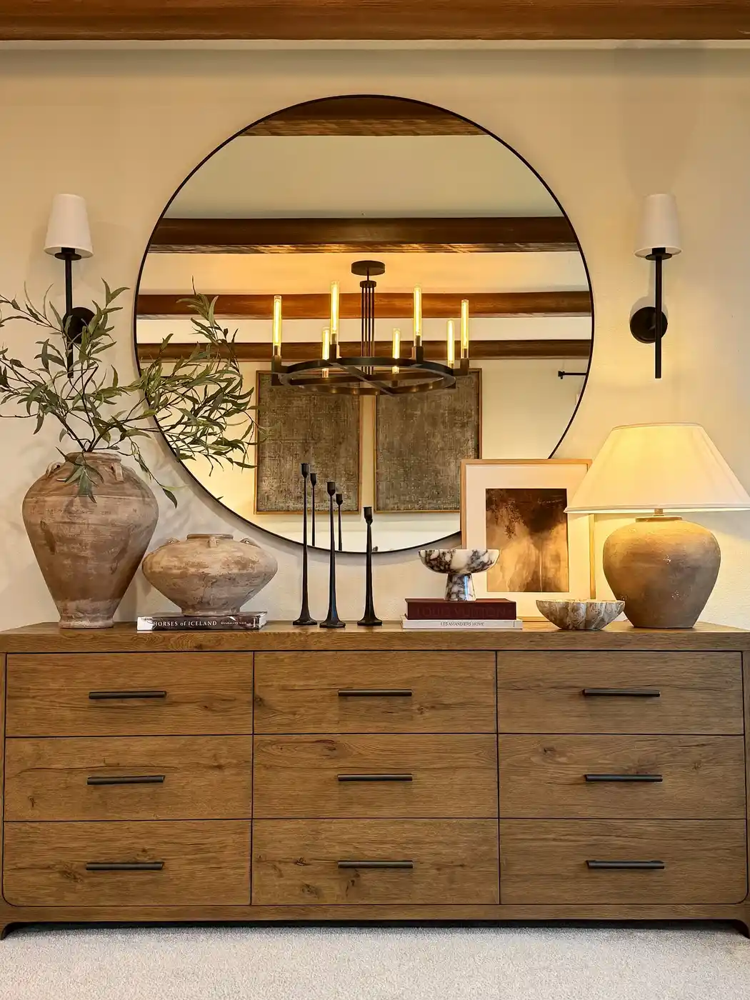 Long wooden dresser beneath oversized mirror with pottery, table lamp, books, and neutral decorative accents