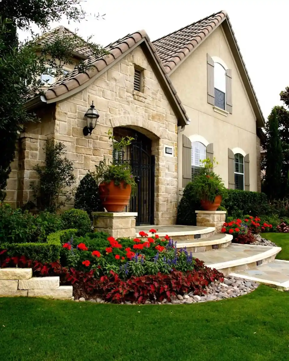 Front entrance surrounded by a low flower bed filled with red flowers beside a stone house