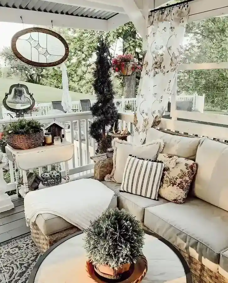 Light neutral covered porch with upholstered seating, patterned pillows, potted plants, and a round wall mirror.